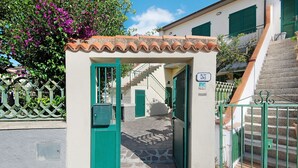 Un estrecho sendero de piedra que conduce a una entrada con portón y un pequeño arco, una escalera a la derecha y un edificio con tejado de tejas y contraventanas verdes.