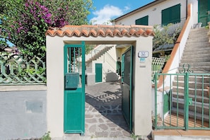 Un Ă©troit sentier de pierre mĂšne Ă une entrĂ©e fermĂ©e par un portail, surmontĂ©e dâune petite arche. Ă droite, on aperçoit un escalier, et en face, un bĂątiment avec un toit en tuiles et des volets verts.