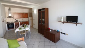 A room with a bed, a dining table with chairs, a kitchen area, a wooden cabinet, and a television on a shelf.