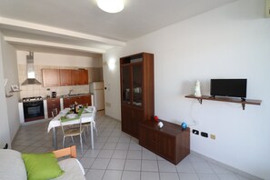 A room with a bed, a dining table with chairs, a kitchen area, a wooden cabinet, and a television on a shelf.