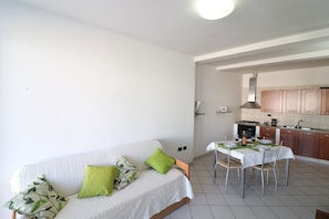 A living room with a sofa and a dining table set for four, a kitchen with wooden cabinets, and a white ceiling.