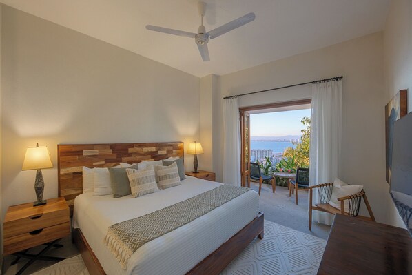 Une chambre d’hôtel équipée d’un lit, de deux tables de chevet, d’un bureau, d’une chaise, d’un ventilateur de plafond et d’un balcon avec vue sur la mer.