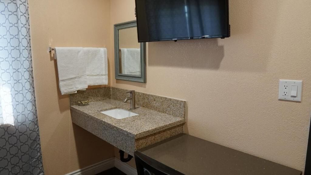 A bathroom with a granite countertop sink, a mirror, a towel rack, and a television mounted on the wall.