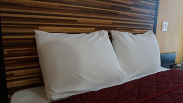 Una cama con almohadas blancas y una colcha roja, frente a un cabecero de madera.