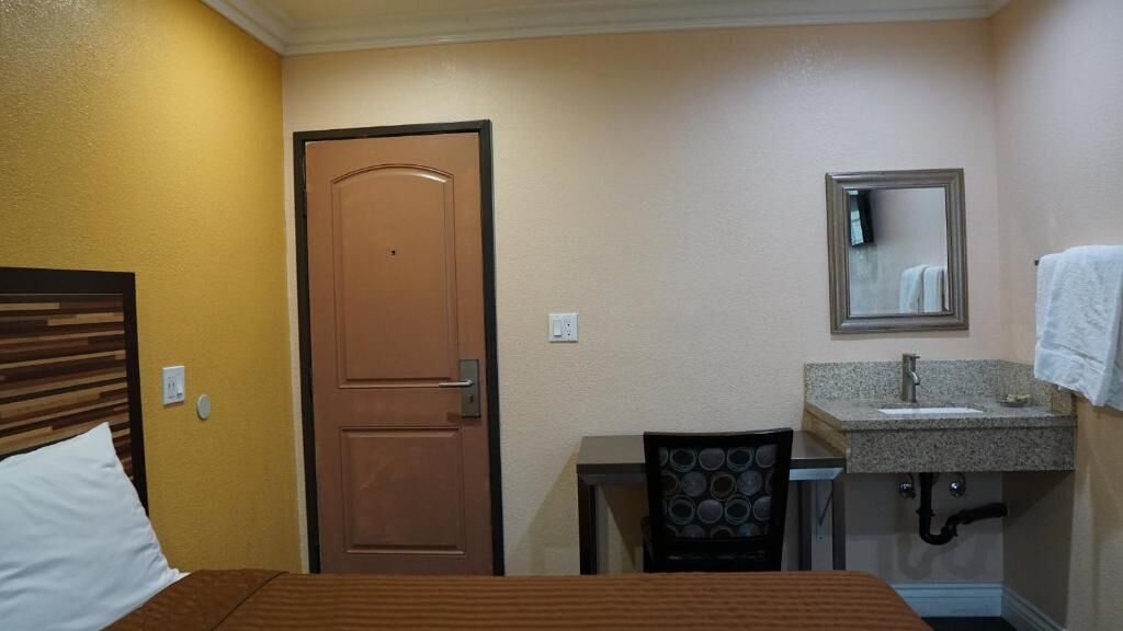 A hotel room with a bed, a wooden door, a small desk with a chair, a sink, and a mirror.