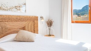 Une chambre avec un grand lit, une tête de lit, une table de chevet ornée d’un vase, une fenêtre avec des rideaux et un luminaire fixé au mur.
