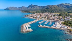 Vue aérienne d’une ville côtière avec un port de plaisance, une digue rocheuse et un paysage montagneux en arrière-plan.