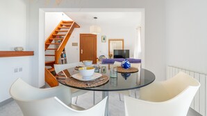 Un espace de repas moderne avec une table en verre, des chaises blanches, un escalier en bois et un salon doté d’un canapé et d’un téléviseur.