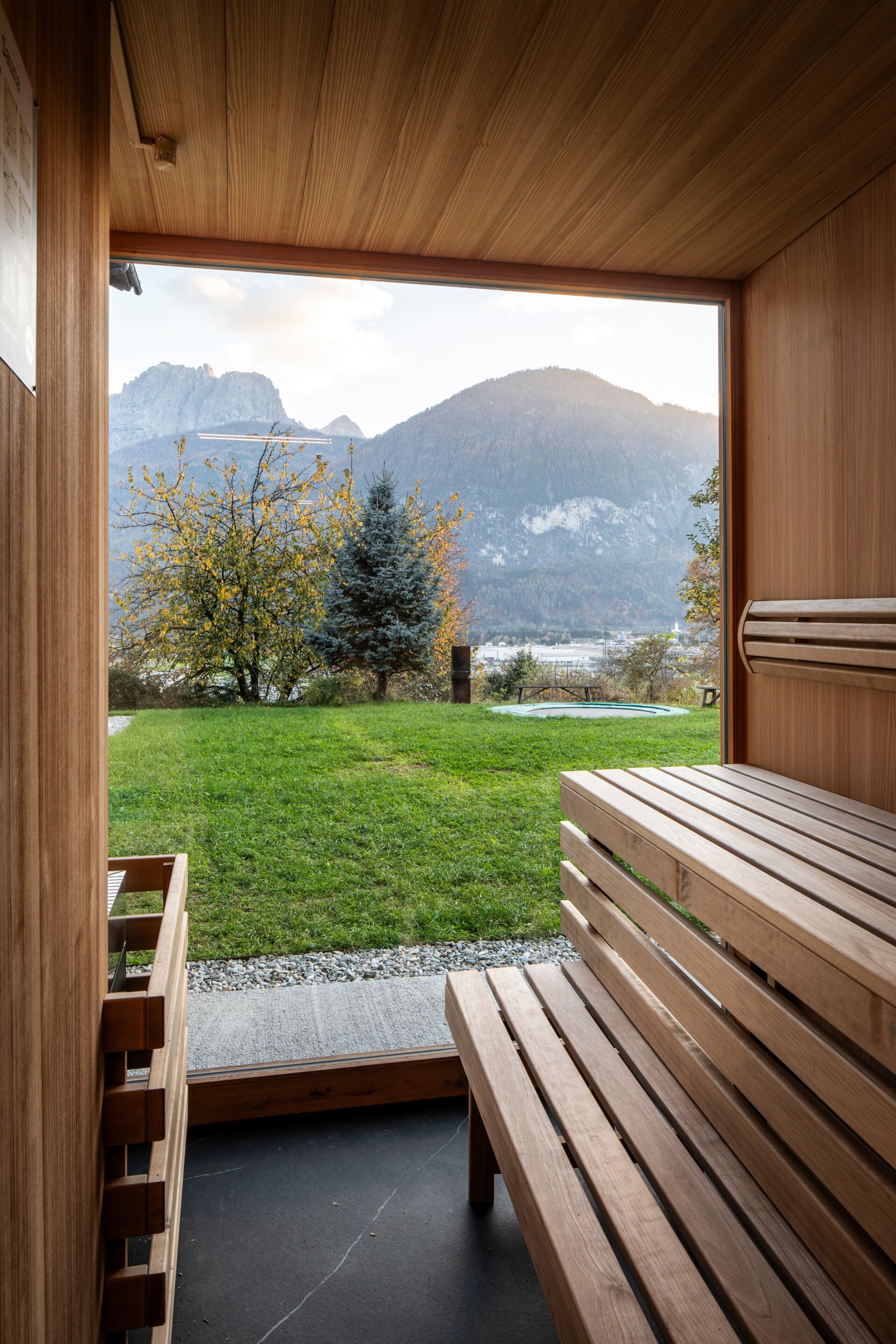 A wooden interior with a bench and a view of a grassy area with trees and mountains.