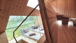 A cabin with a sloped ceiling, wooden walls, and a large window offering a view of a grassy area. There are two beds with pillows and a bench.