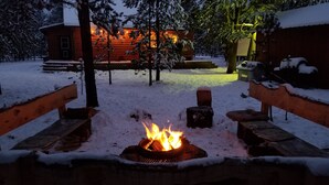 Een besneeuwde buitenruimte met een vuurkuil op de voorgrond, omringd door houten banken.