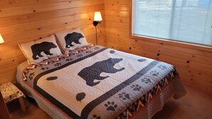 Een gezellige kamer met een houten wand, een bed met quilt met beertjesprint, twee bedlampen en een raam met jaloezieën.