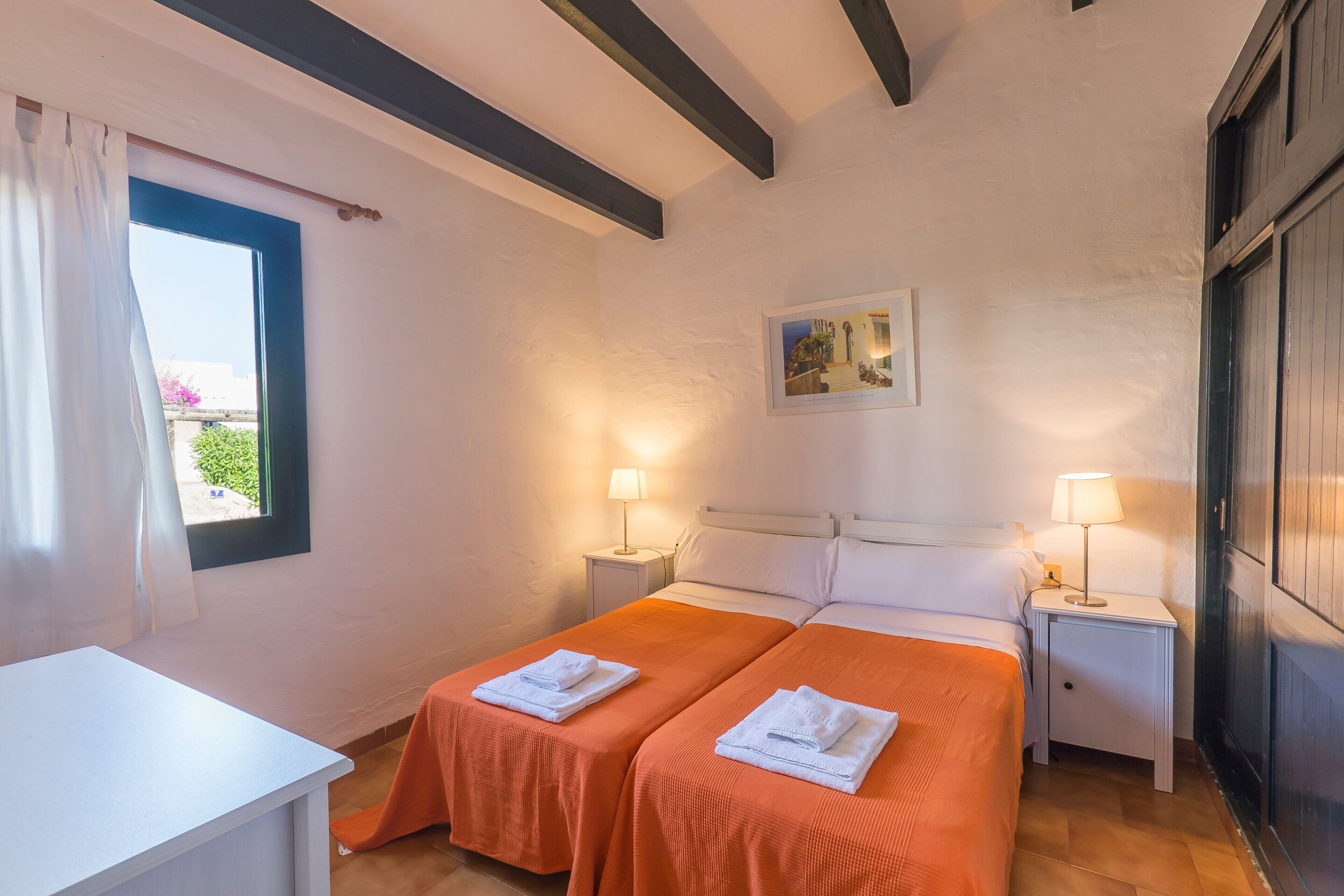 A hotel room with two beds, a desk, a window with curtains, a painting on the wall, and two bedside lamps.