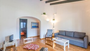 A living room with a grey sofa, a white coffee table, a wooden TV stand, and a painting on the wall. - Beautiful Holiday Home in Residential Complex with Pool, Wi-Fi, Terrace and Lawn (Binisafua)