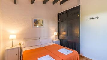 Una cama doble con colcha naranja, una mesita de noche con lámpara, un cuadro en la pared y una puerta oscura.