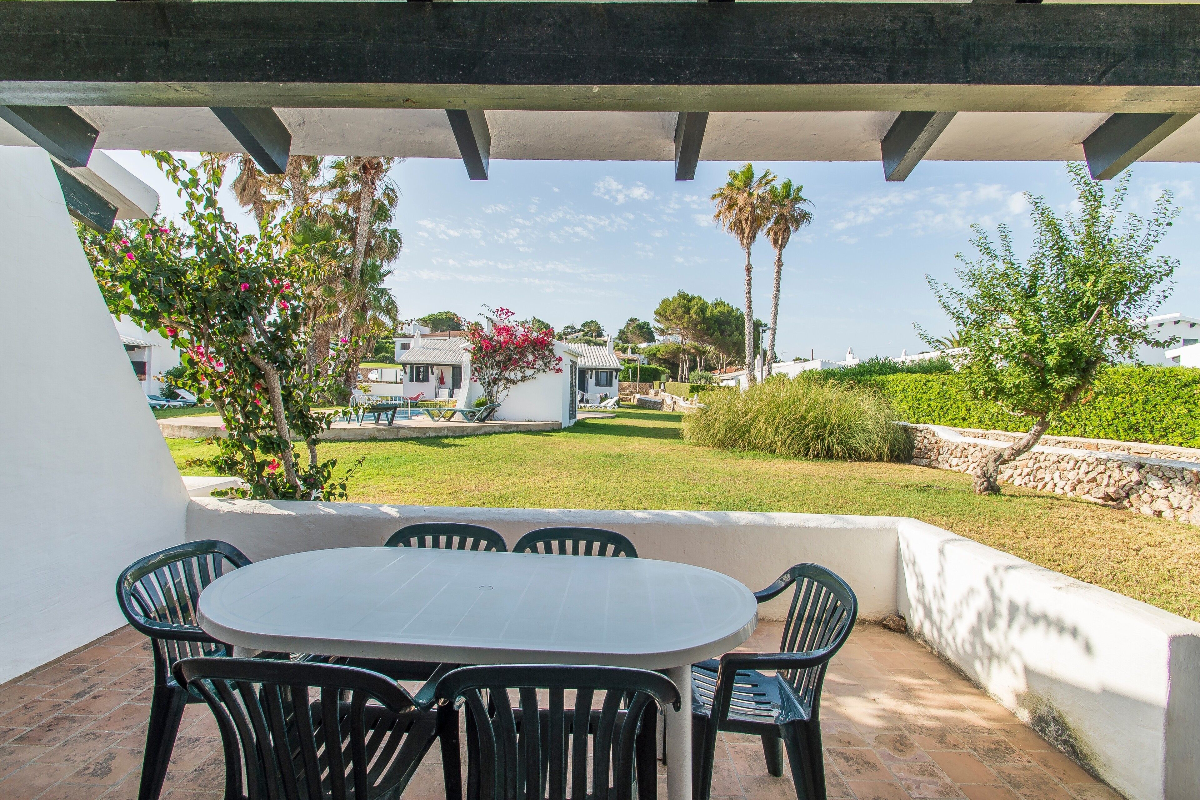 Un patio techado con una mesa y sillas, un muro de piedra y vistas a una zona residencial con casas y vegetación.