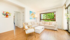 A modern living room with a beige sofa set, a wooden coffee table, and a white armchair. There is a painting on the wall and a window with a view of greenery.