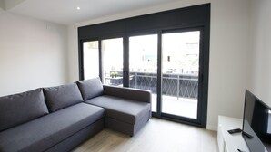 A modern living room with a grey sectional sofa, a flat-screen TV, and a sliding glass door leading to a balcony. - Estudis Laura (Castell-Platja d'Aro)