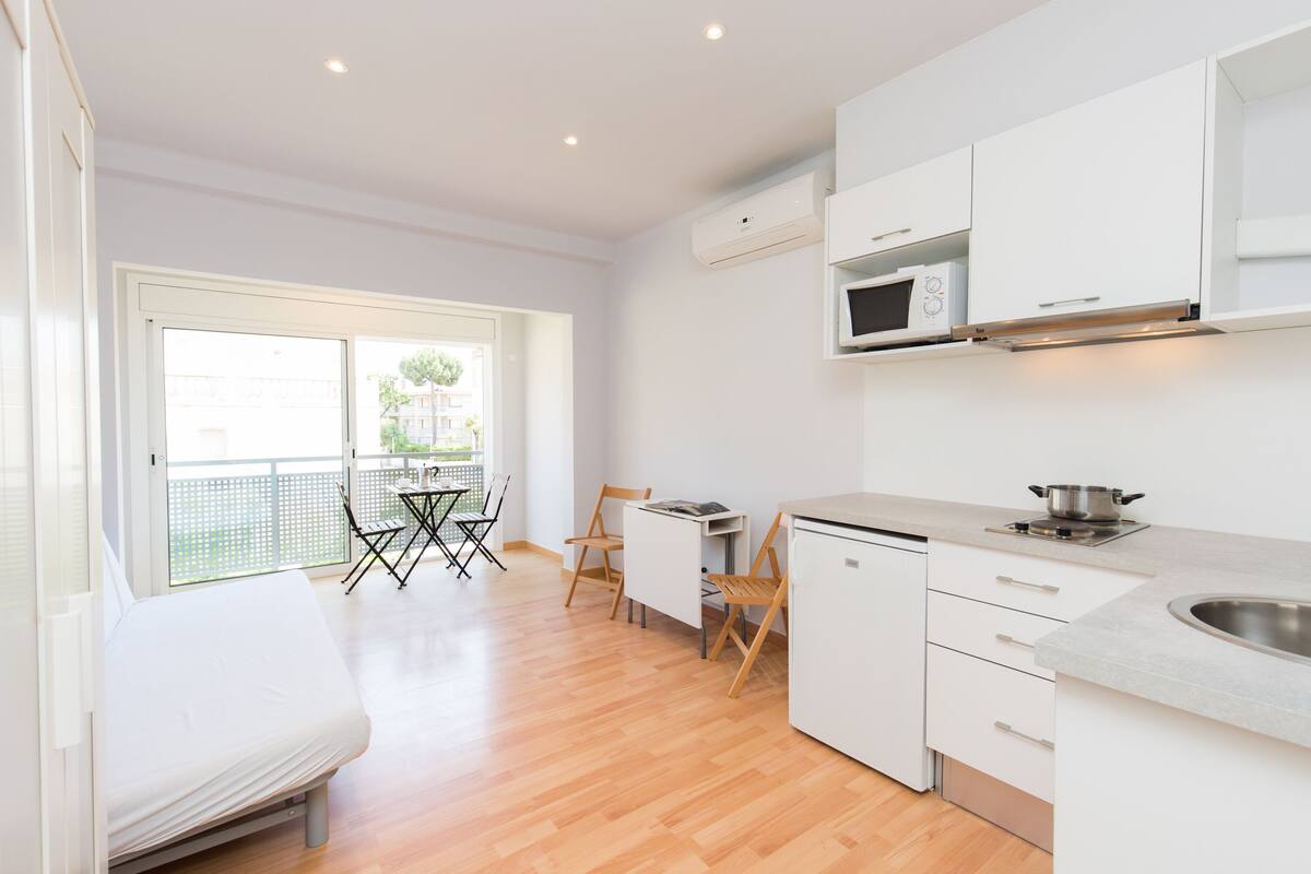 A modern kitchen with white cabinets, a microwave, and a sink. There is a dining area with a table and chairs, and a balcony with a view of the outside.