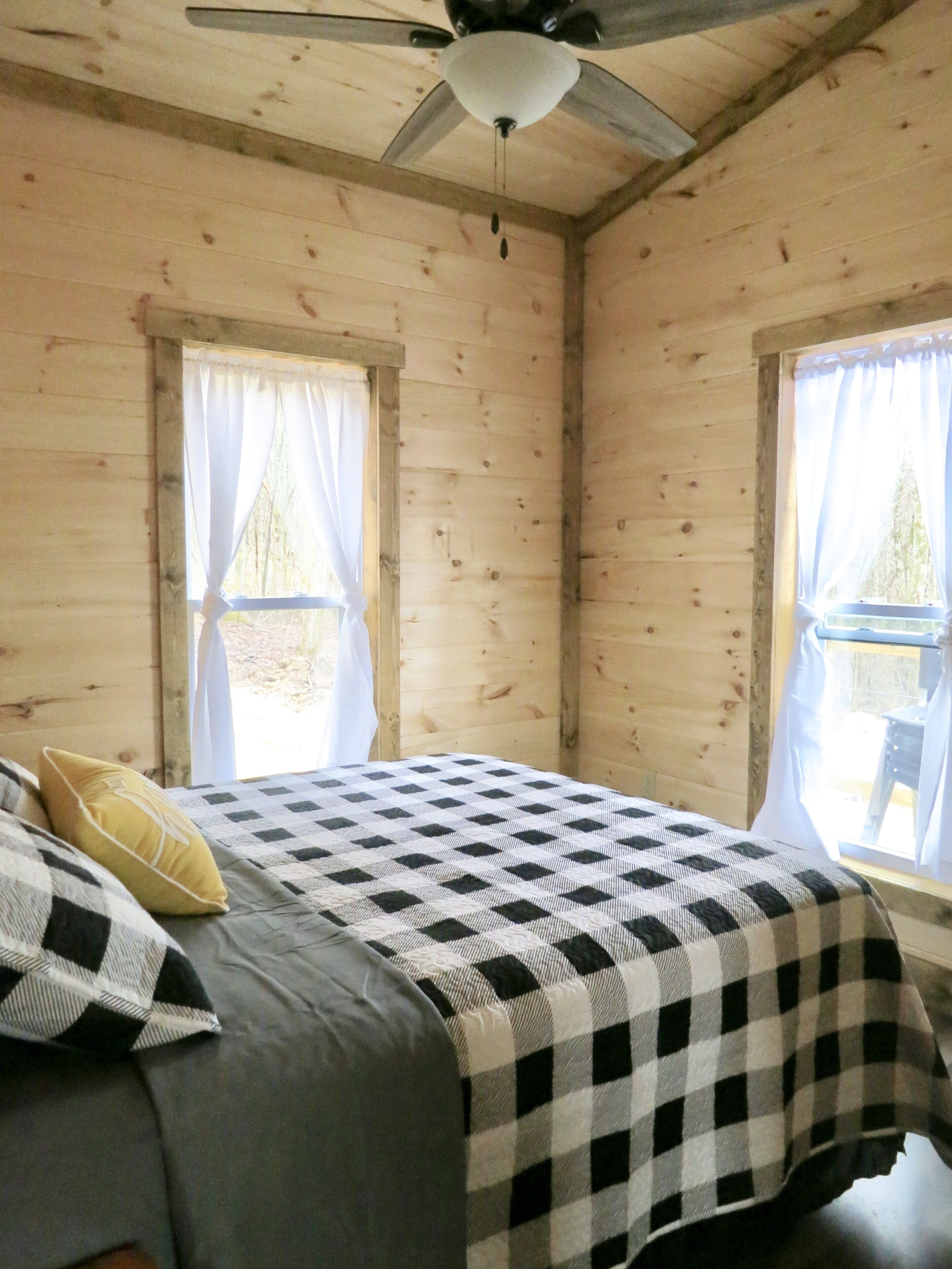 Ein Schlafzimmer einer Holzhütte mit einem Bett, zwei mit Vorhängen versehenen Fenstern und einem Deckenventilator.