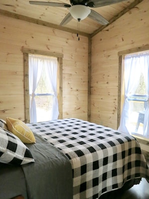 A wooden cabin bedroom with a bed, two windows with curtains, and a ceiling fan.