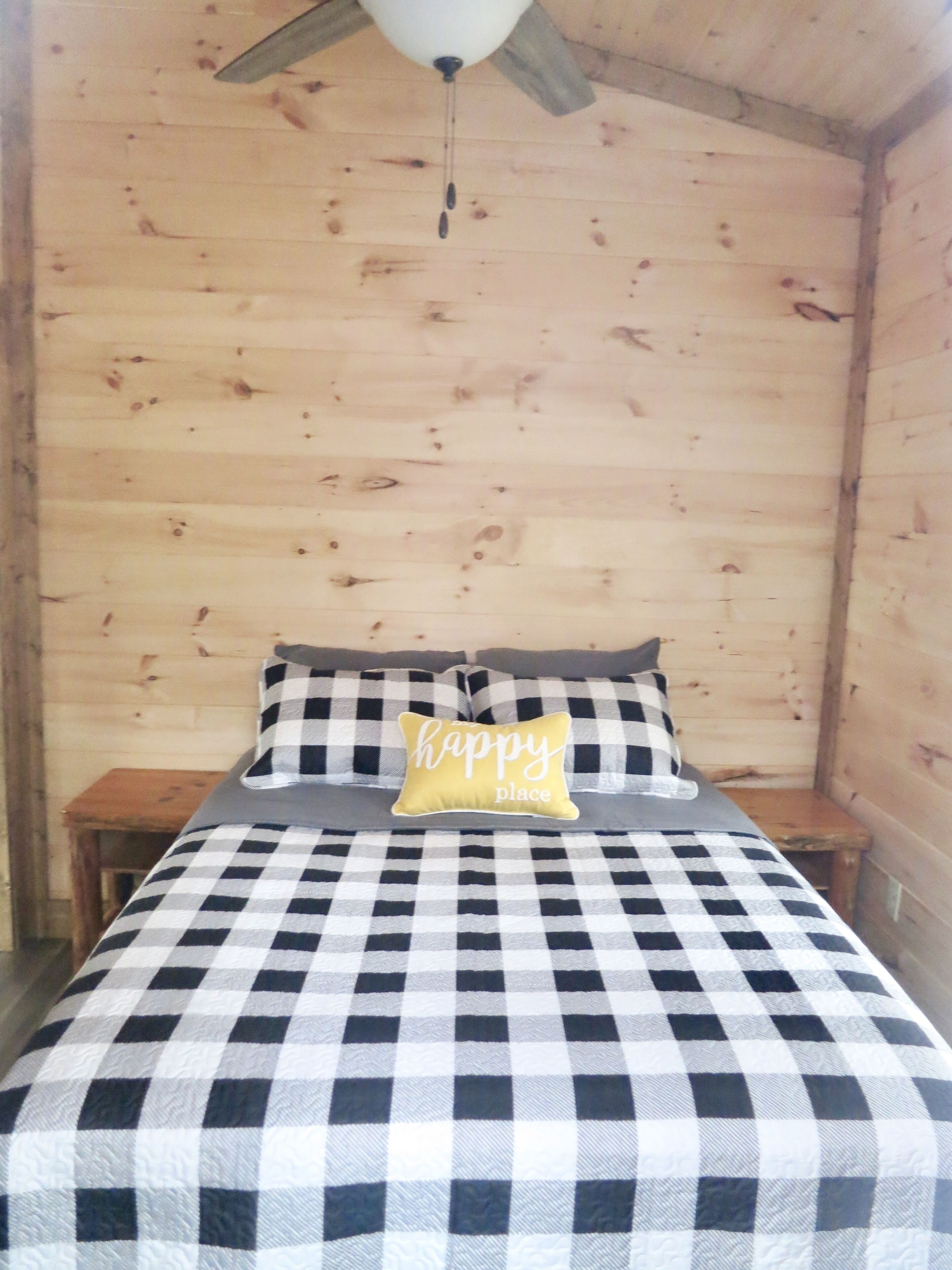 Una habitación con colcha a cuadros, un cojín con la frase 'happy place' y un ventilador de techo.