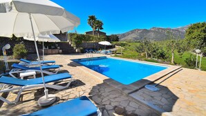 Zona de piscina con sillas reclinables azules y sombrillas blancas, rodeada de baldosas de piedra y follaje, con montañas al fondo.