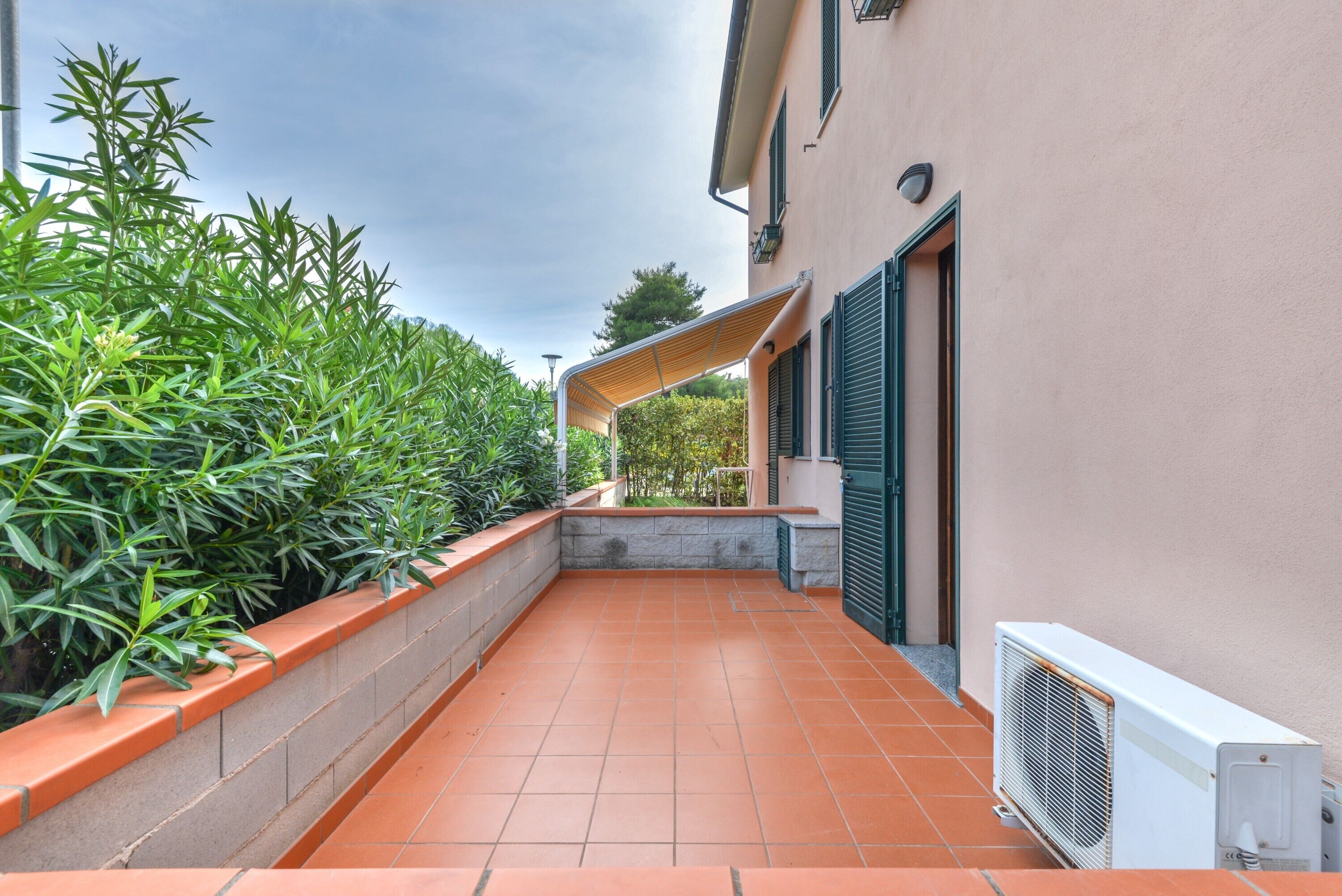 A narrow outdoor corridor with a tiled floor, a green metal railing, and a white air conditioning unit.