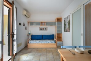 A small, compact room with a bed, a table, and a chair. There is a window with curtains, a wall-mounted air conditioner, and a shelf with decorative items.
