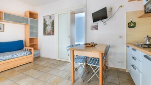A compact living space with a bed, a dining table, chairs, a TV mounted on the wall, and a kitchen area. - Apartment '3B' with Private Terrace and Air Conditioning (Campo nell'Elba)