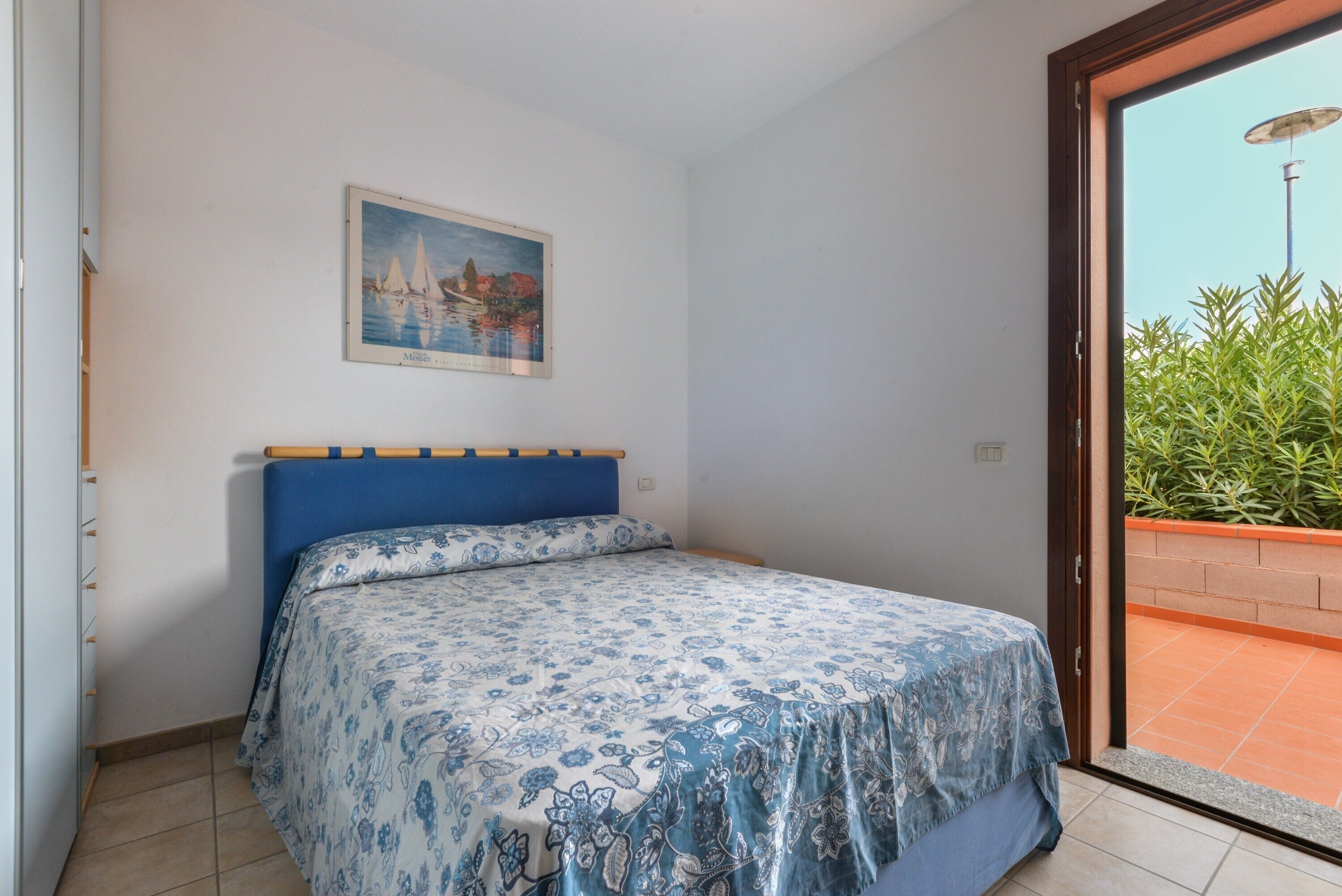 Une chambre avec un lit, un tableau accroché au mur et une vue sur la verdure, visible à travers une porte ouverte.