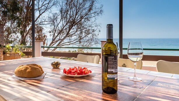Een tafel met een brood, een schaal fruit, een fles wijn en een glas op een terras met uitzicht op de zee.