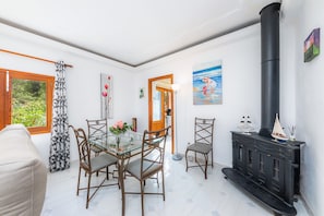 A dining area with a glass table, wooden chairs, a black stove, and a window with a view of greenery.