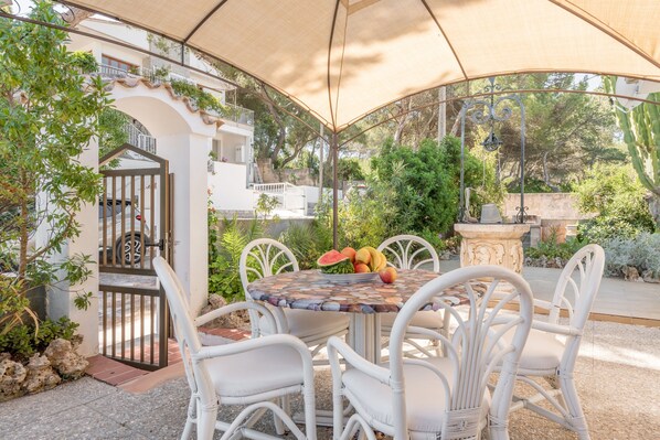 An outdoor patio with a round table, white wicker chairs, and a fruit bowl.