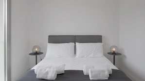 Una cama bien hecha con sábanas blancas, dos lámparas de noche y un cabecero gris.