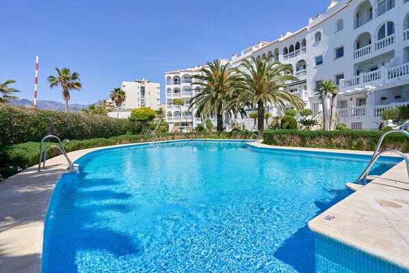 Une piscine à l’eau d’un bleu cristallin, entourée d’un bâtiment blanc doté de balcons et bordée de palmiers.