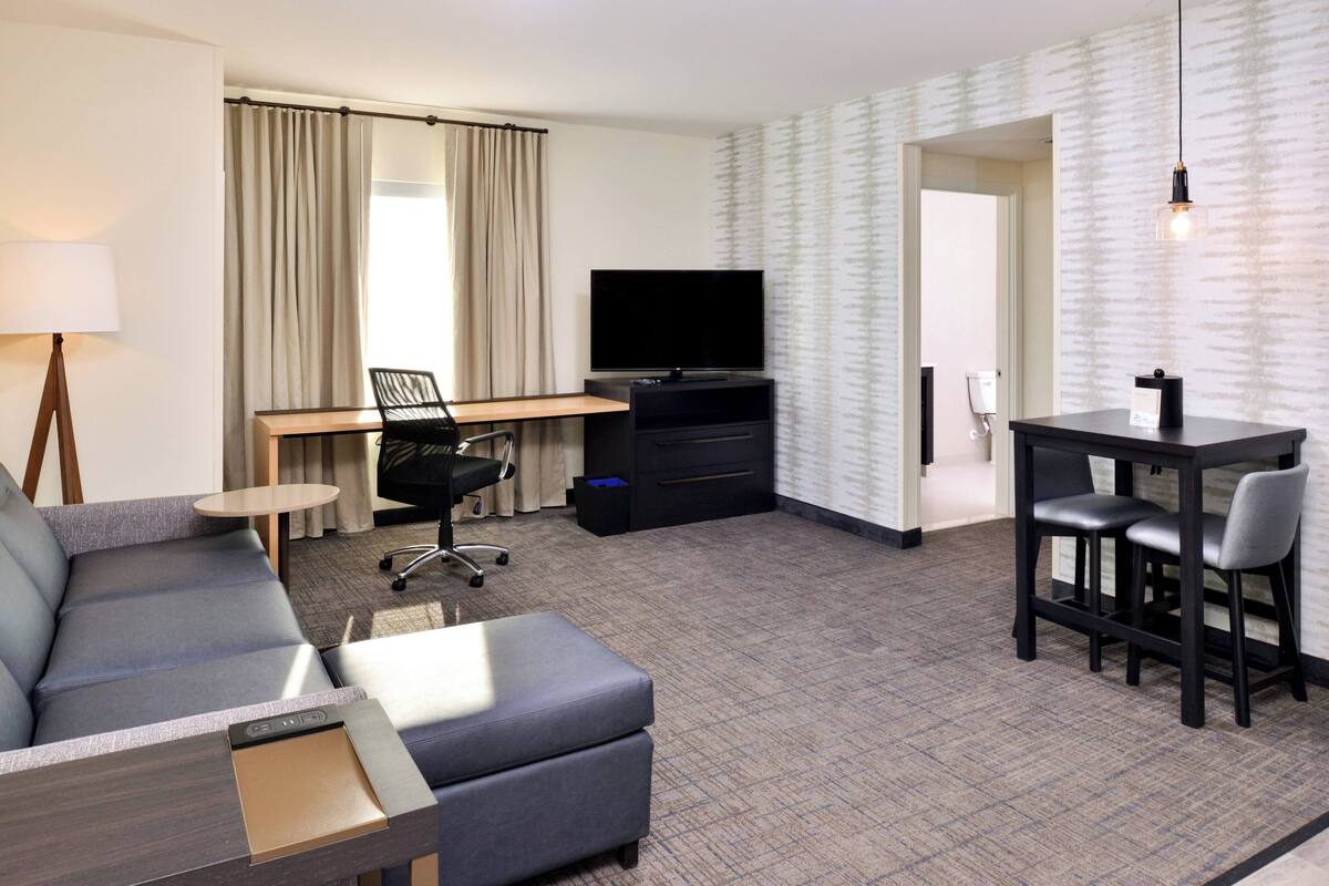 Ein Hotelzimmer mit Sofa, Schreibtisch mit Fernseher, Essbereich mit Stühlen und einer Wand mit Muster.
