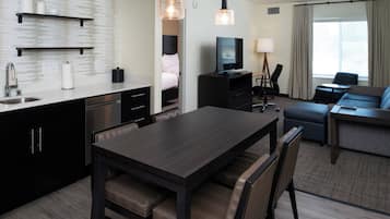 A hotel room with a kitchenette, a dining area, a living area with a sofa and TV, and a large window with curtains.