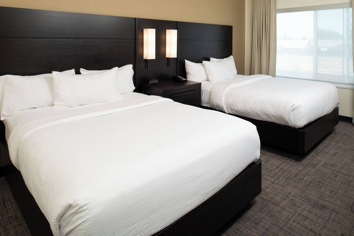 Ein Hotelzimmer mit zwei grossen Betten, einem dunklen Kopfteil, Nachttischlampen und Blick ins Freie durch ein Fenster.