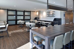 Una sala de conferencias moderna con una amplia mesa de madera, sillas blancas y una cocina con encimera negra y azulejos blancos.