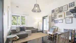 Een moderne woonkamer met een grijze bank, een salontafel en een vloerlamp. Er hangen ingelijste foto’s aan de muur en een raam met witte luiken.