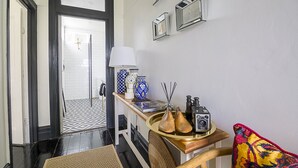 Een gang met een zwart-wit betegelde vloer, een houten bankje met dienblad, een lamp en decoratieve objecten op een tafel.