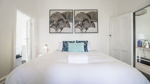 Hotelkamer met een groot bed, twee ingelijste kunstwerken aan de muur, een nachtkastje met een lamp en een deur die leidt naar een andere kamer.