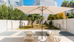Eine Terrasse mit zwei weißen Liegestühlen, einem großen Sonnenschirm und einem kleinen Gartenbereich mit Bäumen und einer Mauer.