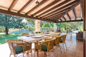 An outdoor patio with a dining table set for four, surrounded by wicker chairs, under a wooden pergola with a view of a swimming pool and greenery.