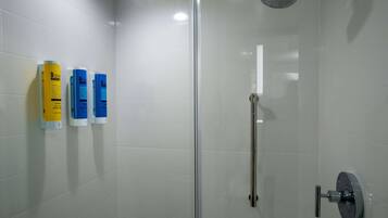 Ducha con cerramiento de vidrio, cabezal de ducha de mano, paredes de azulejos blancos y tres dispensadores de artículos de tocador.