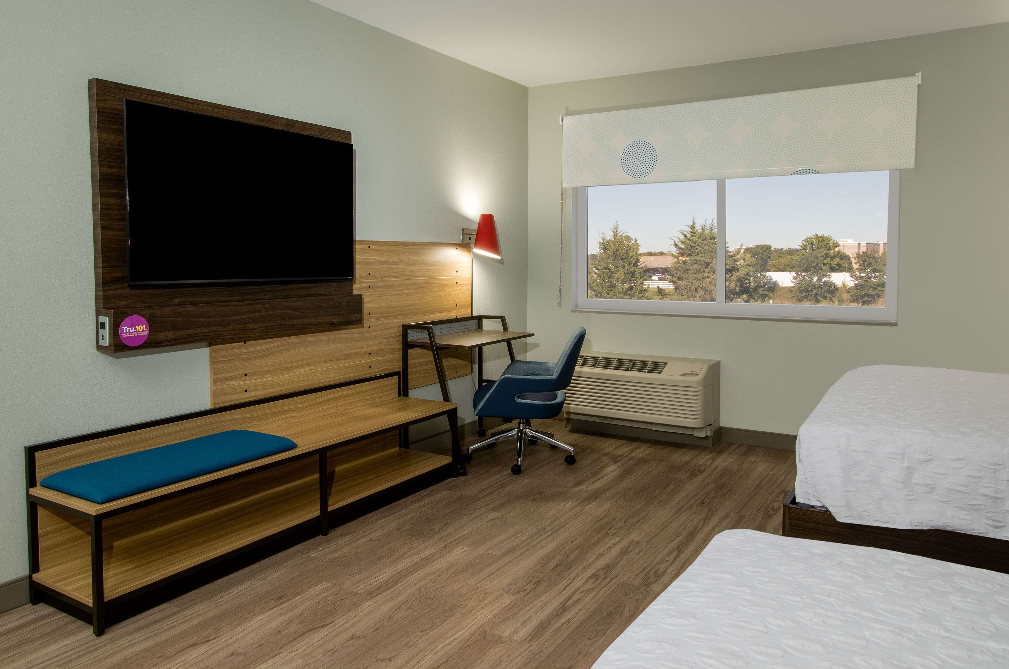 A hotel room with a bed, a desk with a chair, a bench with a cushion, a TV mounted on the wall, and a window with a view of trees and buildings.
