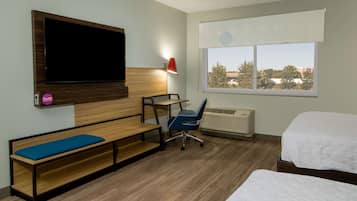 A hotel room with a bed, a desk with a chair, a bench with a cushion, a TV mounted on the wall, and a window with a view of trees and buildings.