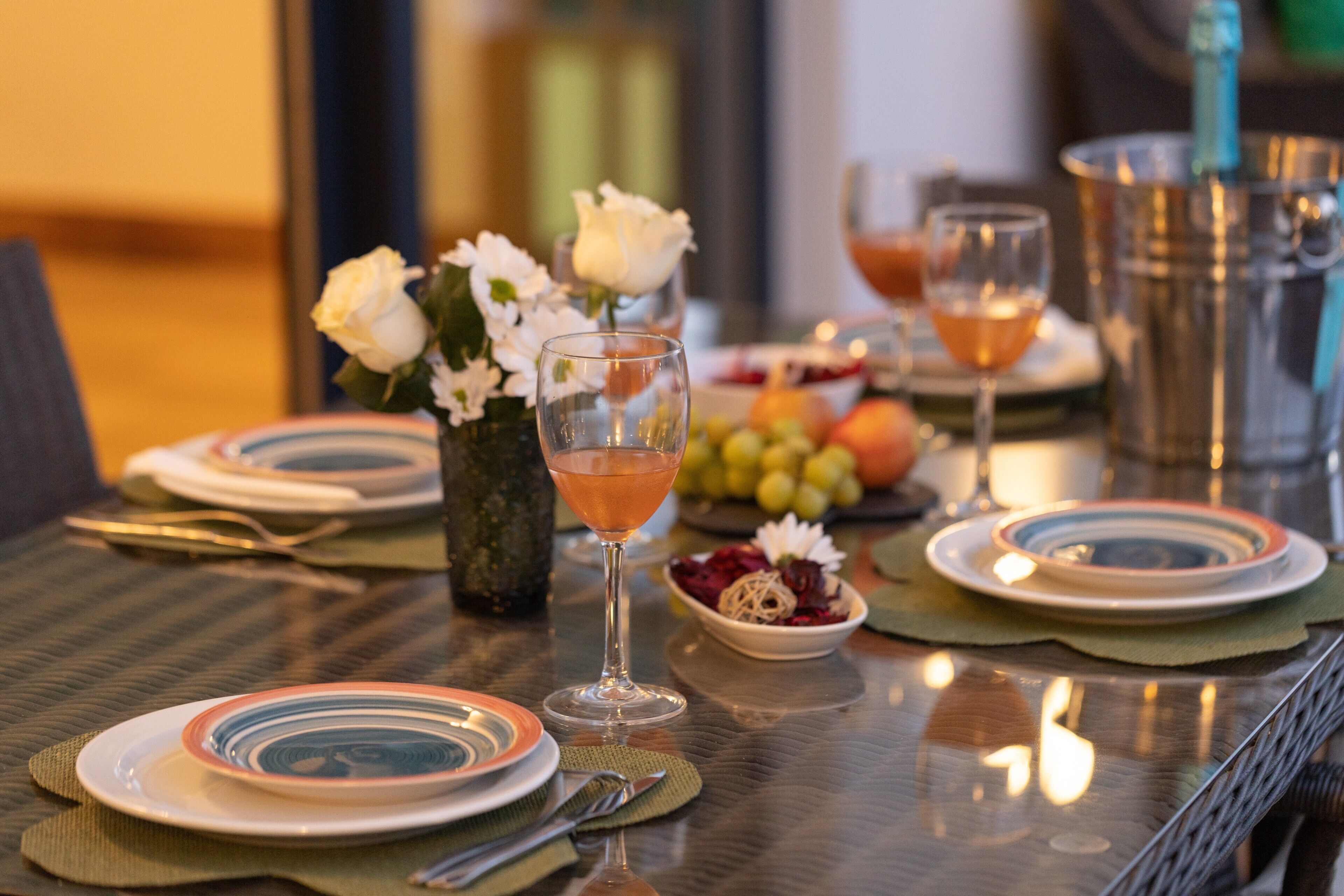Une table à manger dressée avec des assiettes, un verre, un vase de fleurs et un saladier de fruits.