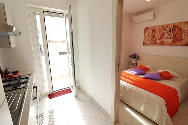 A bedroom with a bed, a painting, and a kitchen area with a stove and cabinets.
